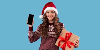 Usmívající se žena ve vánočním oblečení drží telefon a zabalený dárek